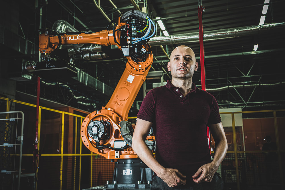 Mujin Corp CEO, Ross Diankov stands in front of a mujin branded orange industrial robot